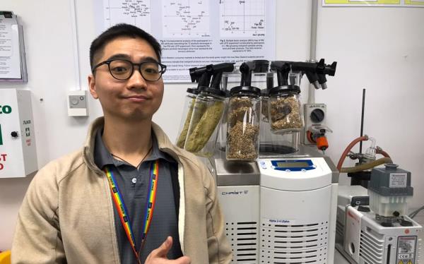 A man wearing glasses, a beige sweater and a rainbow lanyard stands in front of a piece of lab equipment with various different grains in plastic co<em></em>ntainers attached to it. He is smiling and giving a "thumbs up".