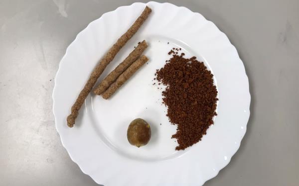 A shot looking straight down o<em></em>nto a white plate which has an avocado seed, a coarse brown powder that looks like coffee grounds, and three twiggy-looking round wafer sticks on it.